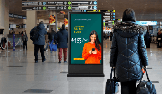 Digital kiosk displaying a mobile carrier advertisement inside a busy airport terminal with travelers walking by.