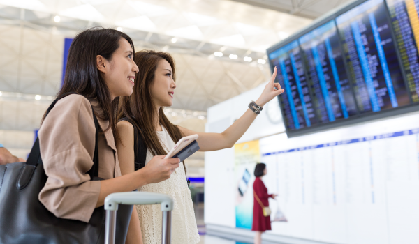 Two travelers looking up at an airport departures board while checking phones, illustrating travel advertising moments and addressable targeting.