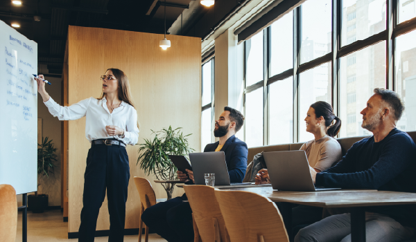 Business professional giving a presentation at a whiteboard to a group of colleagues in a modern office during a B2B strategy meeting.